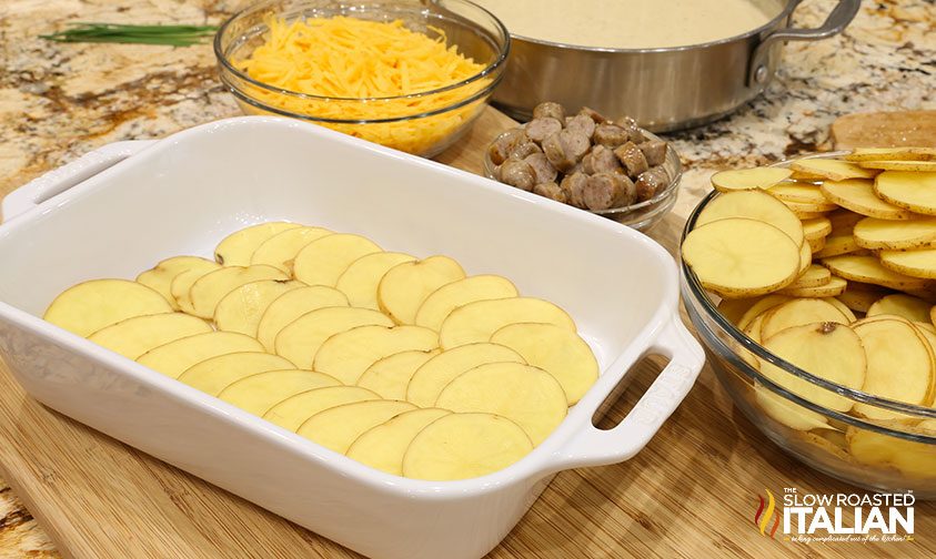 potato slices in casserole dish next to bowls of cheese, sausage, and more potato slices