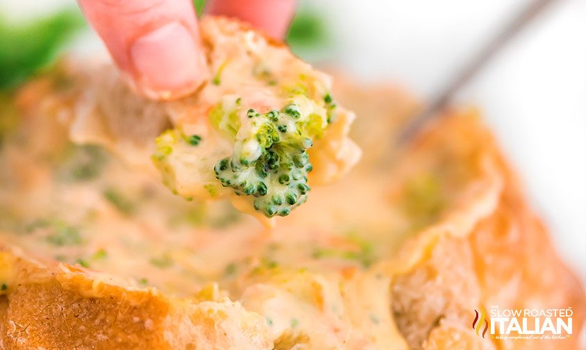 dipping a piece of bread into bread bowl of broccoli cheddar soup