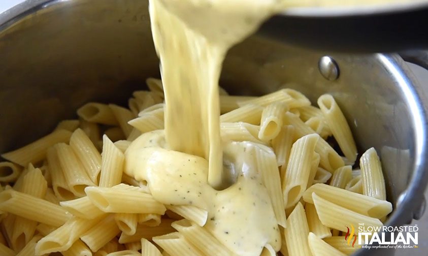 pouring cheese sauce into a pot of cooked penne pasta