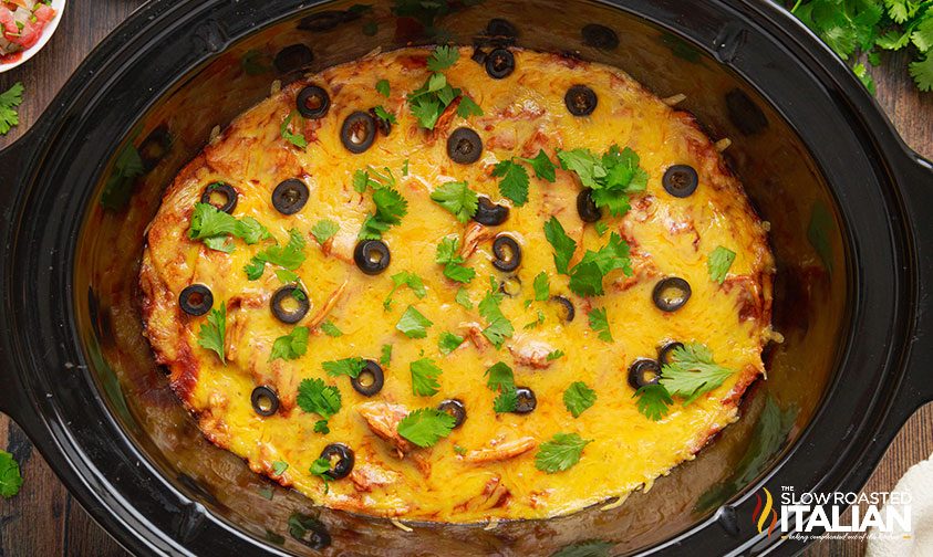 overhead: chicken enchilada casserole in crockpot, topped with cheese, sliced olives, and cilantro