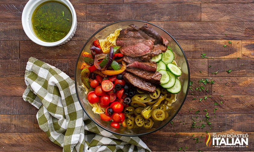 sliced steak and veggies over pasta in a bowl, container of dressing to the side