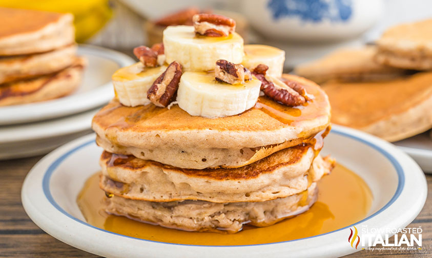 stacked banana nut pancakes drizzled with syrup and slices of banana and pecans on top