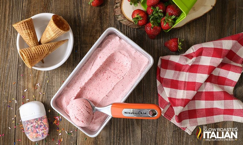 ice cream scoop in pan of homemade strawberry ice cream, surrounded by cones and toppings