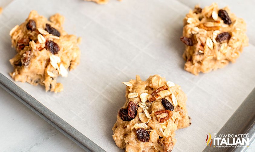 mounds of cookie dough with oats, nuts, and raisins on parchment lined baking sheet