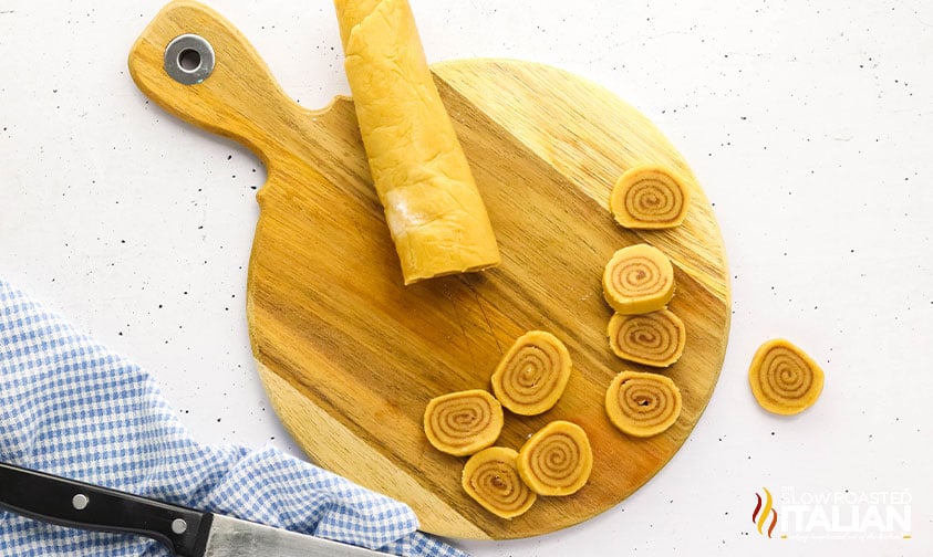 slicing cinnamon roll cookie dough on a wood cutting board