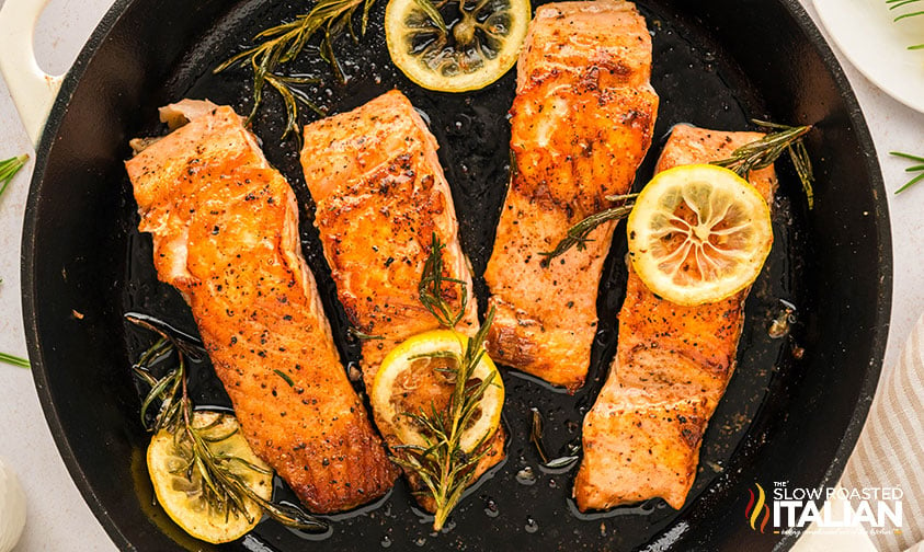 salmon in a cast iron skillet with lemon slices and sprigs of rosemary