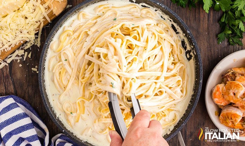 tossing fettuccine noodles in skillet of alfredo sauce with tongs