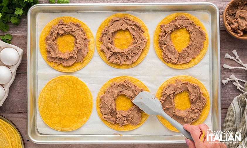 adding refried beans to corn tortillas on a baking sheet