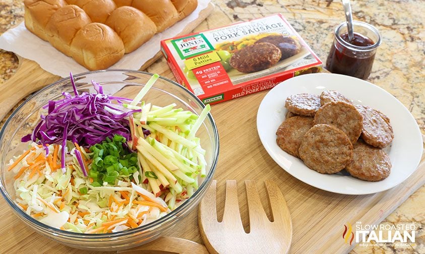 bowl of coleslaw ingredients, plate of cooked sausage patties, and slider buns on a counter