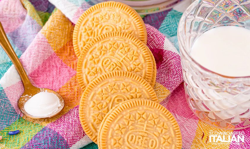 golden Oreos, a glass of milk, and a spoonful of baking powder on a colorful checkered cloth